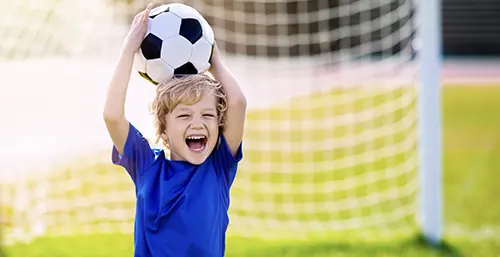 Jakie dyscypliny sportowe są odpowiednie dla przedszkolaków? Mały chłopiec w niebieskiej koszulce i blond włosach podrzuca piłkę do nogi na boisku do piłki nożnej.
