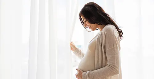 Czy warto stosować probiotyki w ciąży? Szczęśliwa kobieta w ciąży stoi przy oknie przesłoniętym białą firanką i trzyma się za brzuch.